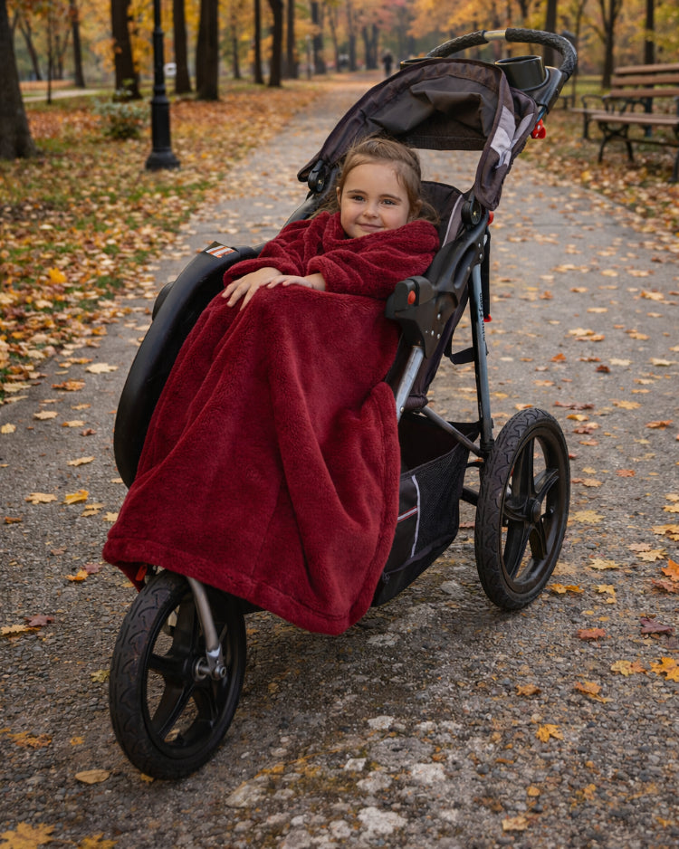 Child in a stroller wrapped in a Ruby Wine Stroller Slanket on a leaf-covered path