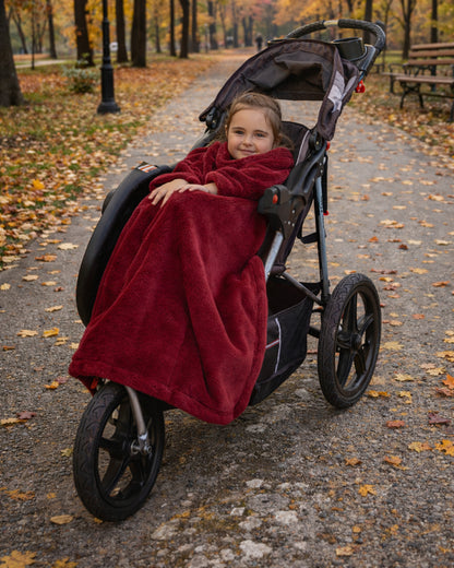 Child in a stroller wrapped in a Ruby Wine Stroller Slanket on a leaf-covered path