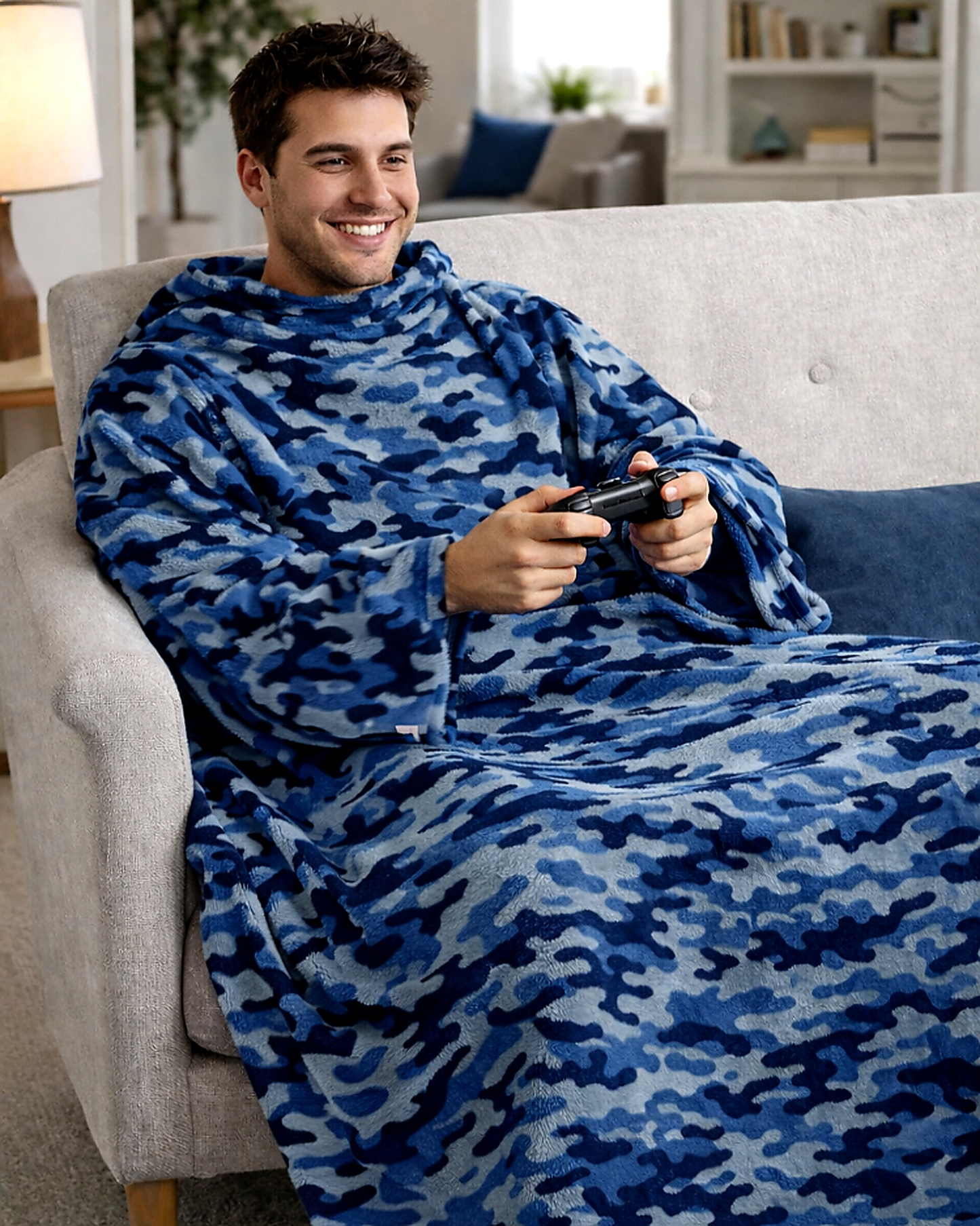 Man sitting on a couch wearing a blue camouflage blanket, holding a gaming controller.